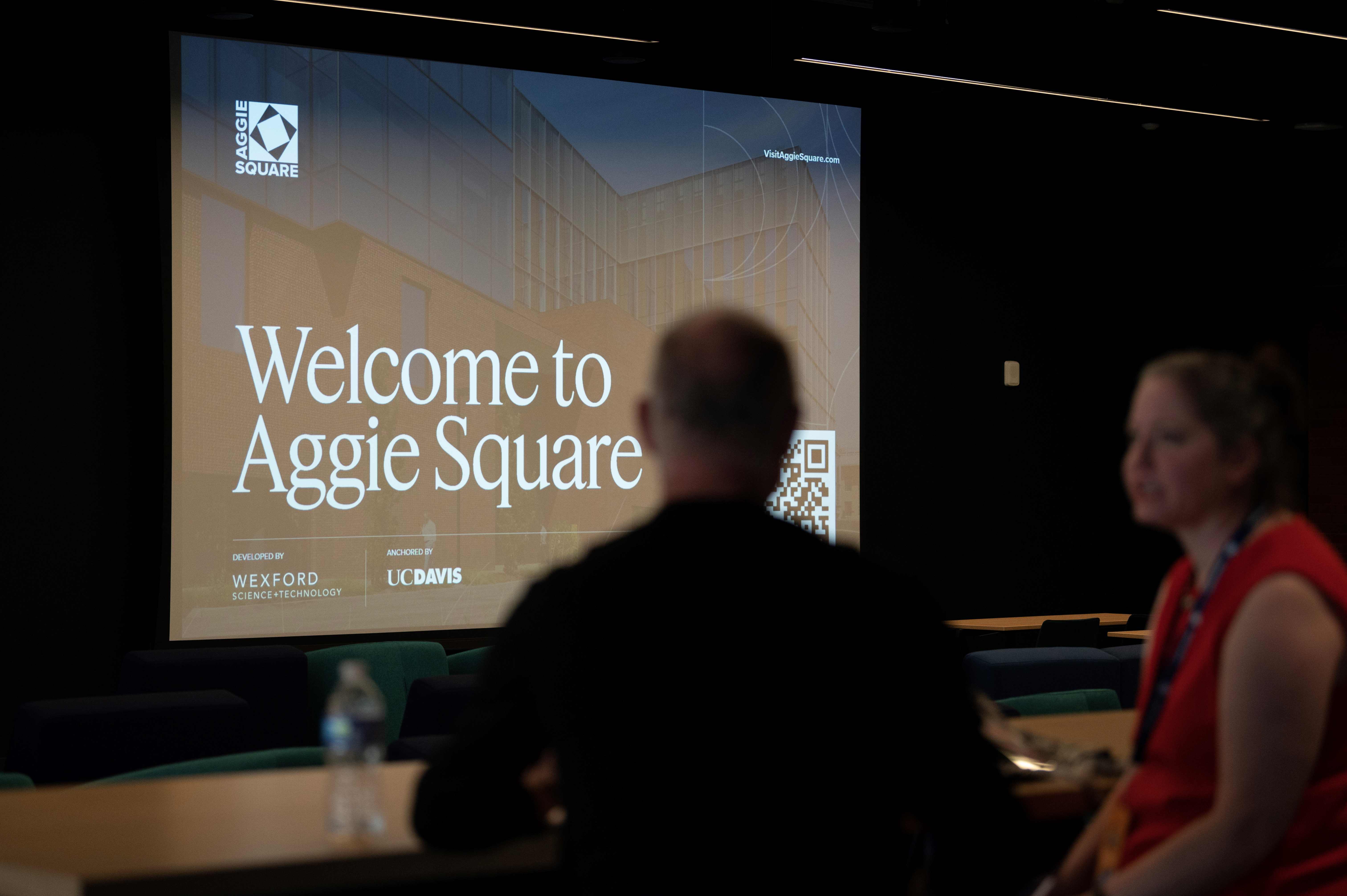 A man and a woman chat in the foreground of the picture, with a large screen saying "welcome to Aggie Square" in the background.