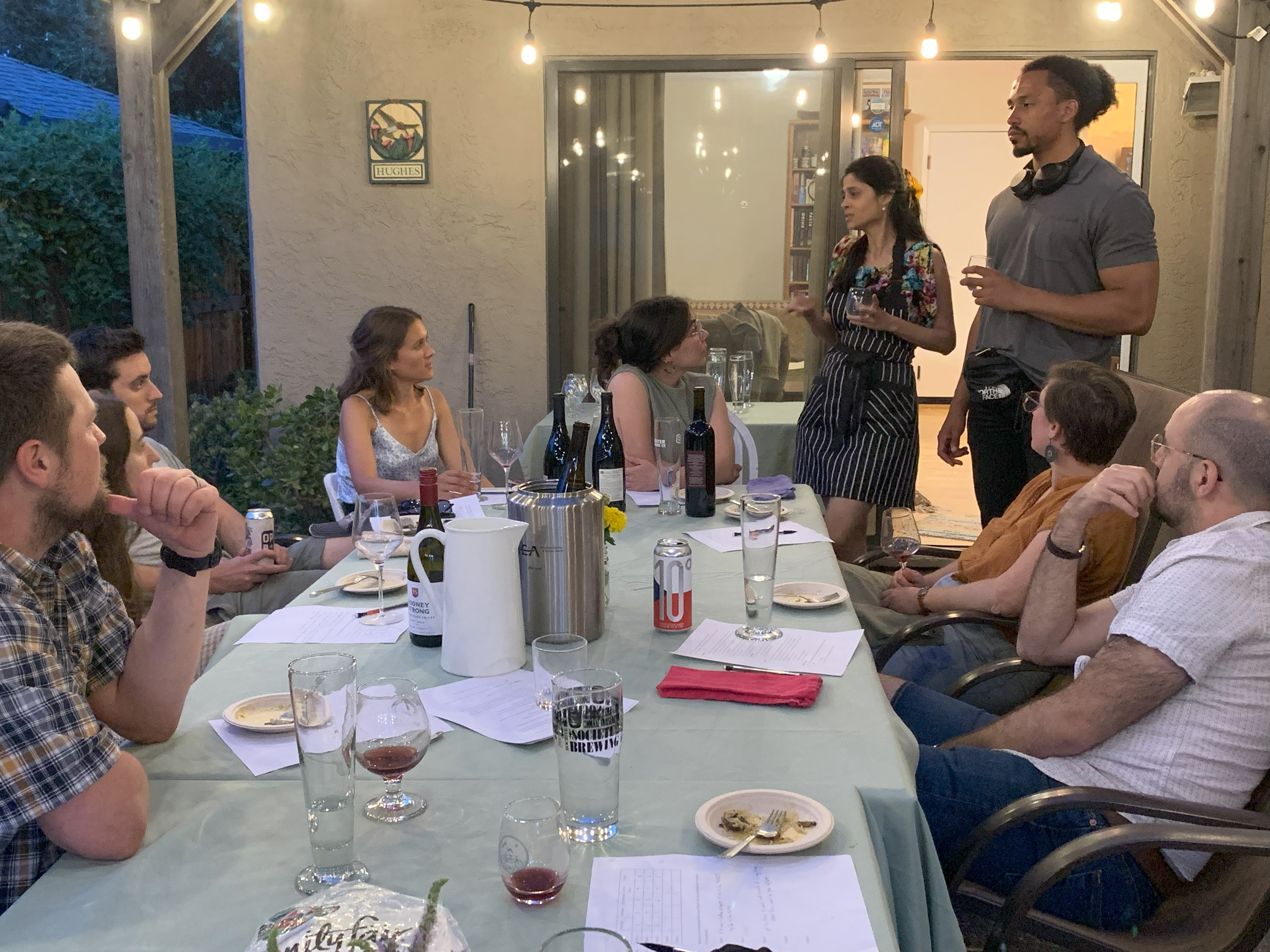 A group of people sit around a table covered in glasses, plates and notes. The seated people are all looking at a man and a woman who are standing up at the end of the table, addressing the group.