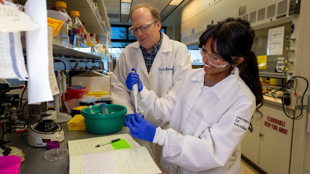 A man and a woman wearing lab coats work at a lab bench.