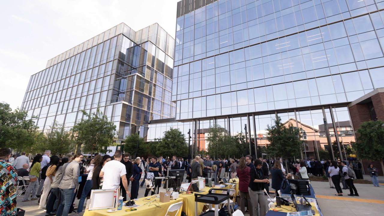 Hundreds of people mingling outside a glass and brick building with tables set up showcasing different organizations.