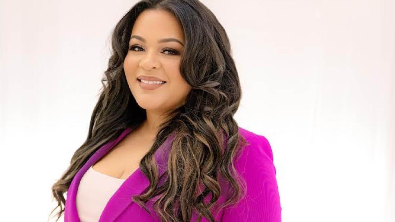 An African American woman wearing a pink jacket smiles towards the camera.