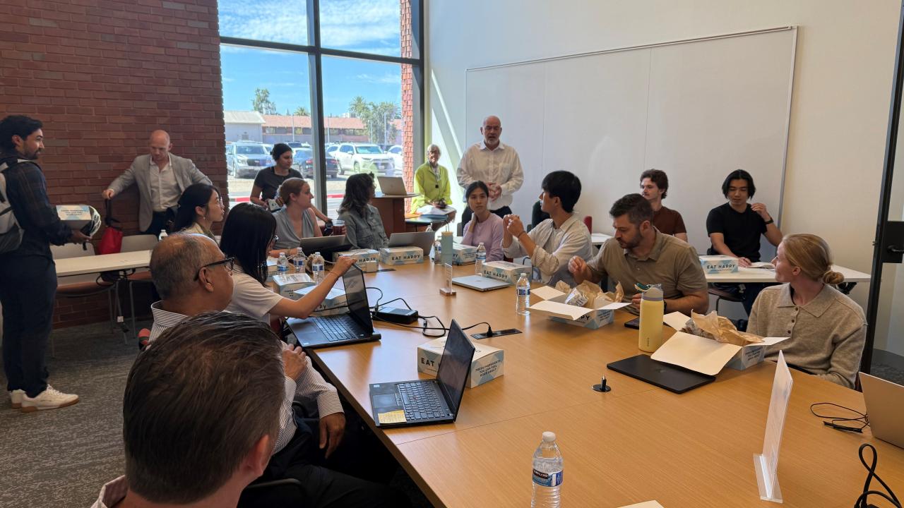 A group of people sit around a large conference room desk, listening as a man in a white shirt stands and addresses the audience.