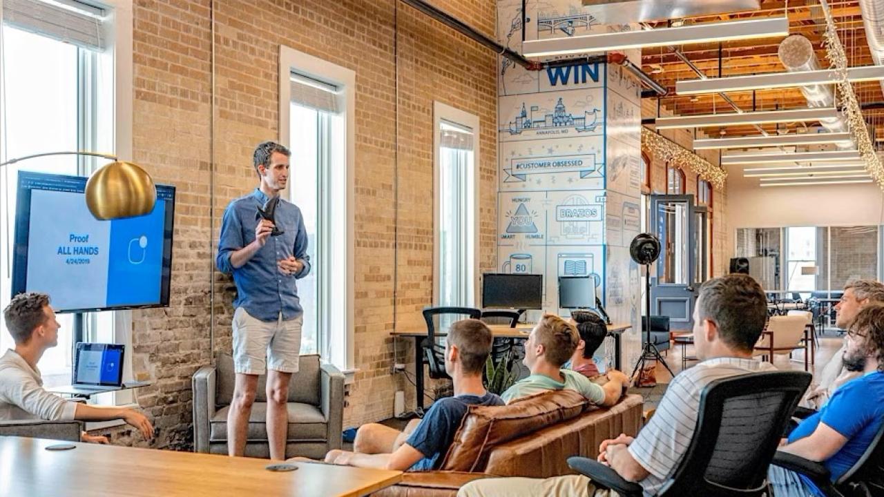 People sitting on couches and chairs in a modern office space listening to a man in a blue shirt and shorts give a presentation