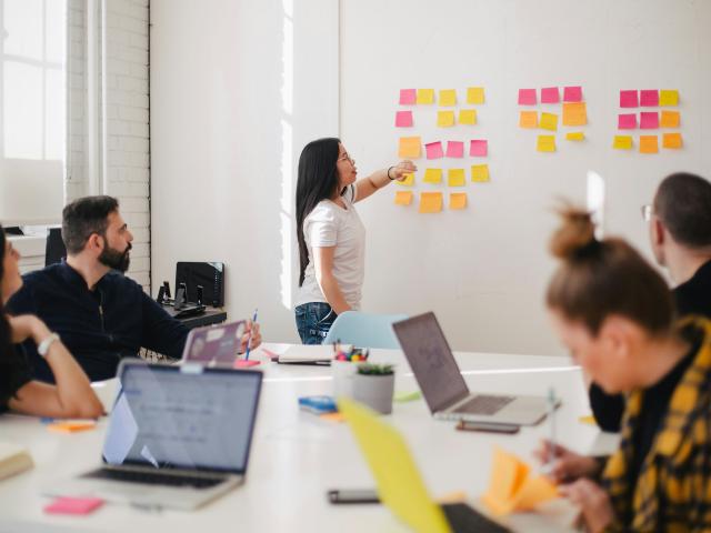 Woman stands at white board pointing at post-it notes while coworkers at a conference table listen.