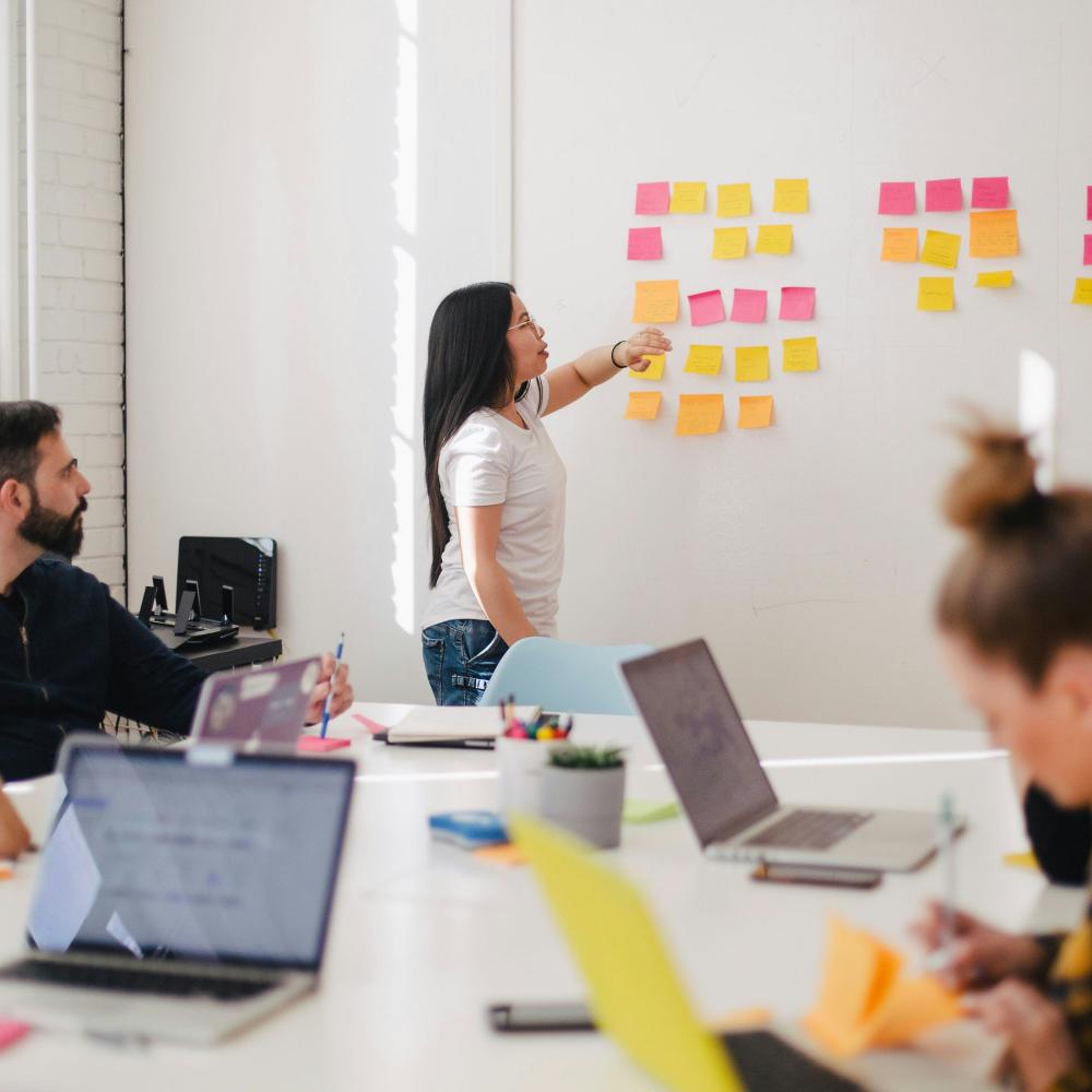 Woman stands at white board pointing at post-it notes while coworkers at a conference table listen.