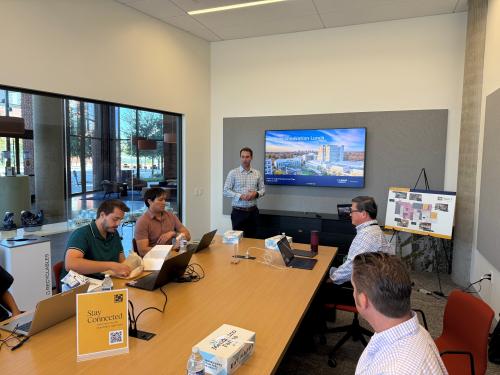 A man stands at the front of a conference room, speaking in front of a display screen as people seated at a conference room table listen.