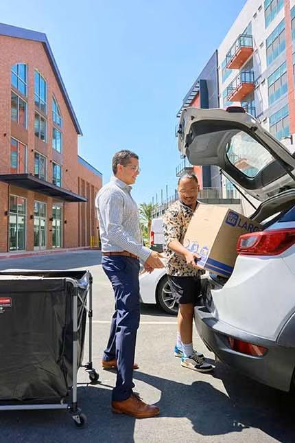 A man in slacks and a dress shirt assists another man with a cardboard box he's lifting out of the trunk.