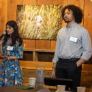 A man and a woman stand in front of a wood-paneled wall. The man looks on while the woman speaks into a microphone.