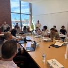 A group of people sit around a large conference room desk, listening as a man in a white shirt stands and addresses the audience.