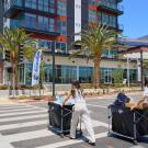 Two women in beige clothing push carts with belongings across the street towards the ANOVA Aggie Square apartment, a new building with a brick facade and lots of windows. 