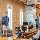 People sitting on couches and chairs in a modern office space listening to a man in a blue shirt and shorts give a presentation